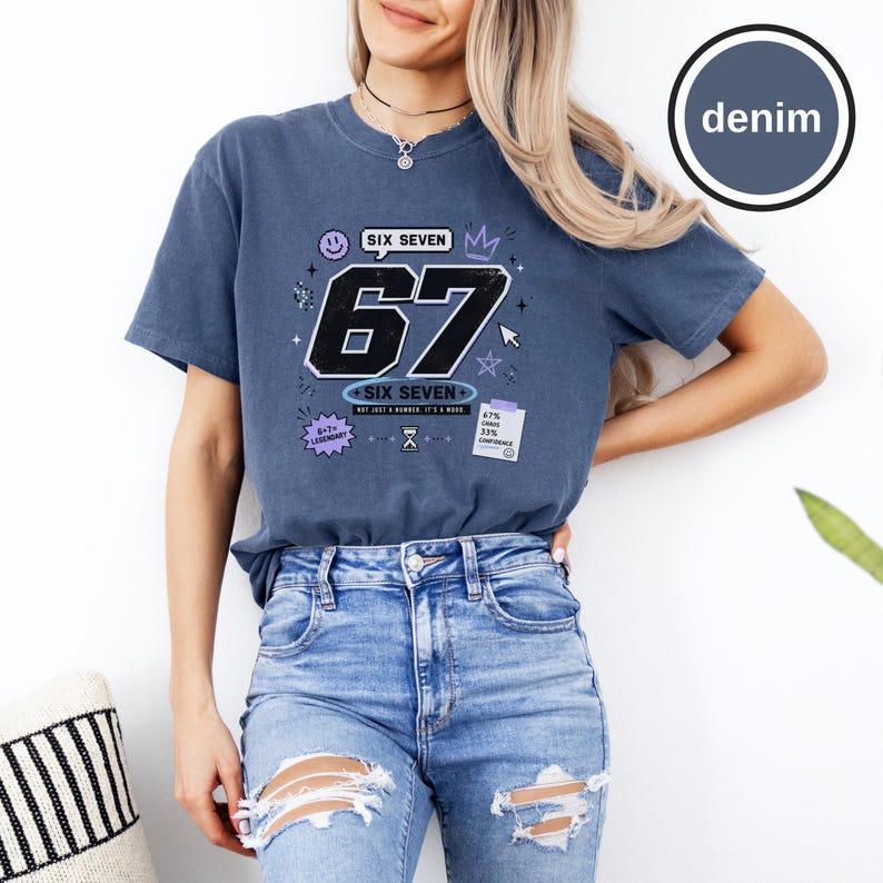 Pu&ograve; includere: Una t-shirt blu denim con una grande grafica del numero 67 in nero con contorni bianchi. Il design include il testo SIX SEVEN, una faccina sorridente, una corona, un cursore e una piccola nota con il testo 67% CHANCE 33% CONFIDENCE.