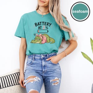 May include: A seafoam green short-sleeve t-shirt featuring a graphic of a tired green alligator wearing a pink headband. Above the alligator, the text reads BATTERY AT 2% with a low battery icon. The shirt has a crew neckline and a relaxed fit.