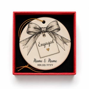 May include: A round ceramic ornament in a red gift box, featuring a black and white sketch of a bow and a tag with the text Engaged and a small gold heart. Below the tag, the text reads Name & Name and MM.DD.YYYY. A gold string is attached.