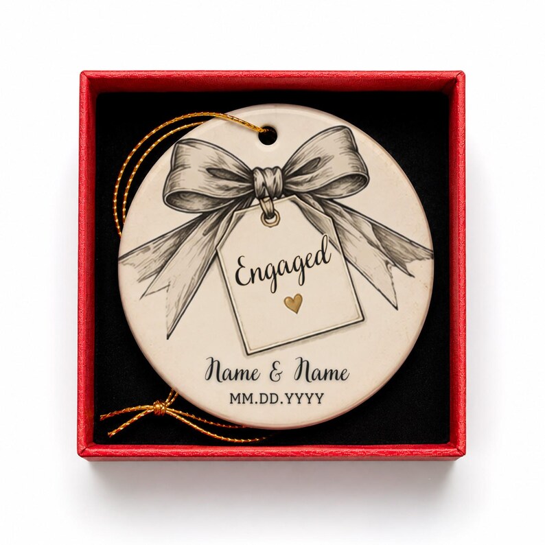 May include: A round ceramic ornament in a red gift box, featuring a black and white sketch of a bow and a tag with the text Engaged and a small gold heart. Below the tag, the text reads Name & Name and MM.DD.YYYY. A gold string is attached.