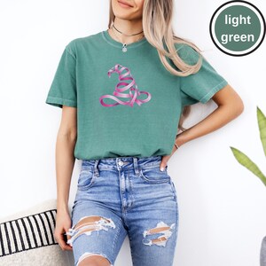May include: A light green short-sleeve t-shirt featuring a graphic of a witch hat constructed from a pink awareness ribbon. The shirt has a crew neckline and a relaxed fit. A circular badge in the top right corner contains the text light green.