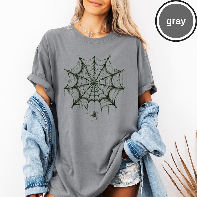 May include: A gray short-sleeve t-shirt featuring a dark green spiderweb graphic centered on the chest with a small spider hanging from the bottom of the web. The word gray is printed in a white circle in the upper right corner of the image.