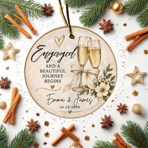 K&ouml;nnte beinhalten: Ein runder Keramikschmuck mit zwei Champagnergl&auml;sern, einer beigen Schleife, Blumenmotiven und dem Text Engaged and a beautiful journey begins, Emma & James, 25.10.2026. Das Design enth&auml;lt goldene Herzen und eine goldene Aufh&auml;ngeschnur.