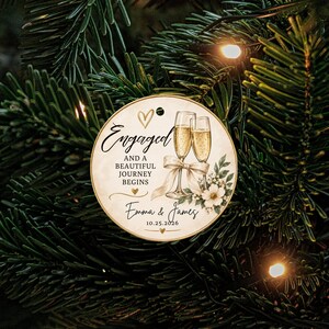 K&ouml;nnte beinhalten: Ein runder, cremefarbener Anh&auml;nger mit zwei Champagnergl&auml;sern, einer wei&szlig;en Schleife, Blumenmotiven und dem Text: Engaged AND A BEAUTIFUL JOURNEY BEGINS, Emma & James, 10.25.2026, sowie kleinen goldenen Herzsymbolen.