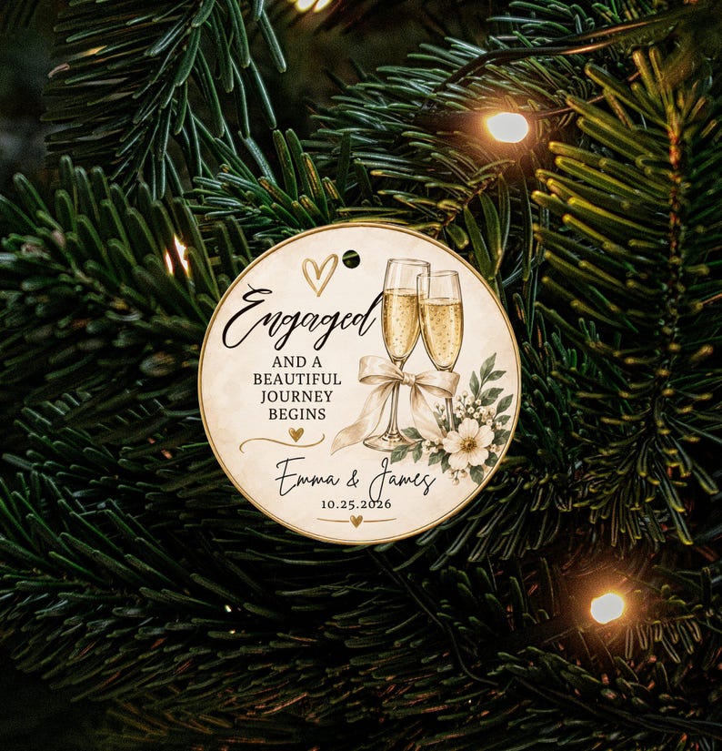 K&ouml;nnte beinhalten: Ein runder, cremefarbener Anh&auml;nger mit zwei Champagnergl&auml;sern, einer wei&szlig;en Schleife, Blumenmotiven und dem Text: Engaged AND A BEAUTIFUL JOURNEY BEGINS, Emma & James, 10.25.2026, sowie kleinen goldenen Herzsymbolen.