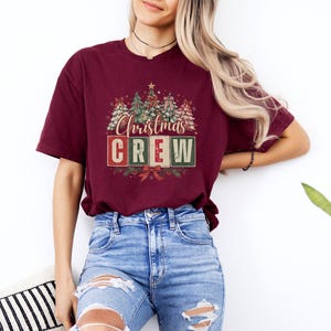 Op de afbeelding: Een bordeauxrood t-shirt met korte mouwen en een grafische print van gestileerde kerstbomen en de tekst "Christmas CREW" in rode en groene blokletters, geaccentueerd met een klein sterretje bovenaan en een rode strik onderaan.