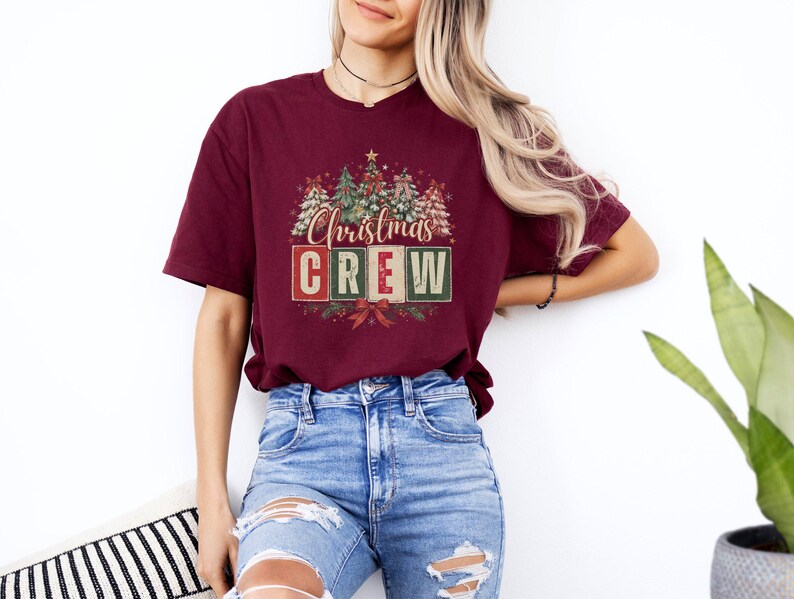 Op de afbeelding: Een bordeauxrood t-shirt met korte mouwen en een grafische print van gestileerde kerstbomen en de tekst "Christmas CREW" in rode en groene blokletters, geaccentueerd met een klein sterretje bovenaan en een rode strik onderaan.