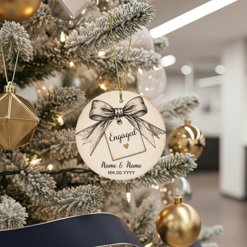 May include: A round, cream-colored ceramic ornament hanging by a gold string. The design features a black line-art illustration of a bow tied to a gift tag with the word Engaged, a small gold heart, and placeholders for Name & Name and MM.DD.YYYY.