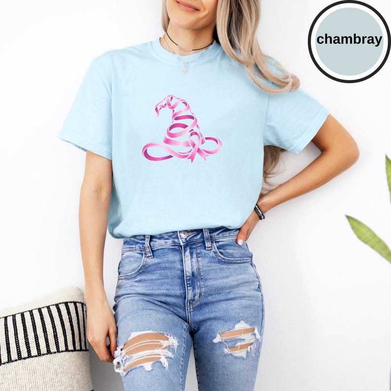 May include: A light blue short-sleeve t-shirt featuring a graphic of a witch hat constructed from a pink awareness ribbon. The shirt is labeled as chambray. The garment has a crew neckline and a relaxed fit, paired with distressed blue denim jeans.