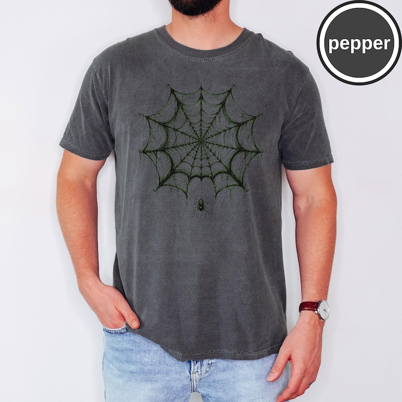 May include: A charcoal gray crew neck t-shirt featuring a dark green spiderweb graphic centered on the chest with a small spider hanging from the bottom of the web. The word pepper is printed in white text inside a black circle in the top right corner.