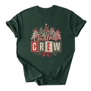 Op de afbeelding: Een donkergroen T-shirt met korte mouwen en een grafische print van vijf versierde kerstbomen met rode strikken en sterren. Onder de bomen staat de tekst "Christmas CREW" in een gestileerd lettertype, met het woord CREW in vier blokken.