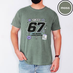 Pu&ograve; includere: Una t-shirt a maniche corte color verde muschio con una stampa grafica che presenta il grande numero nero 67, il testo SIX SEVEN e varie piccole icone tra cui una faccina sorridente, una corona, una clessidra e una nota con 67% CHAOS 33% CONFIDENCE.