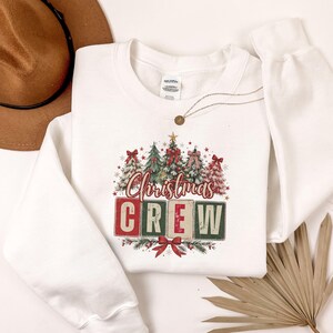 Op de afbeelding: Een wit sweatshirt met lange mouwen en een afbeelding van vier gestileerde kerstbomen versierd met rode strikken en sterren. Onder de bomen staat de tekst "Christmas CREW" in rode en groene blokletters met een rode strik onderaan.