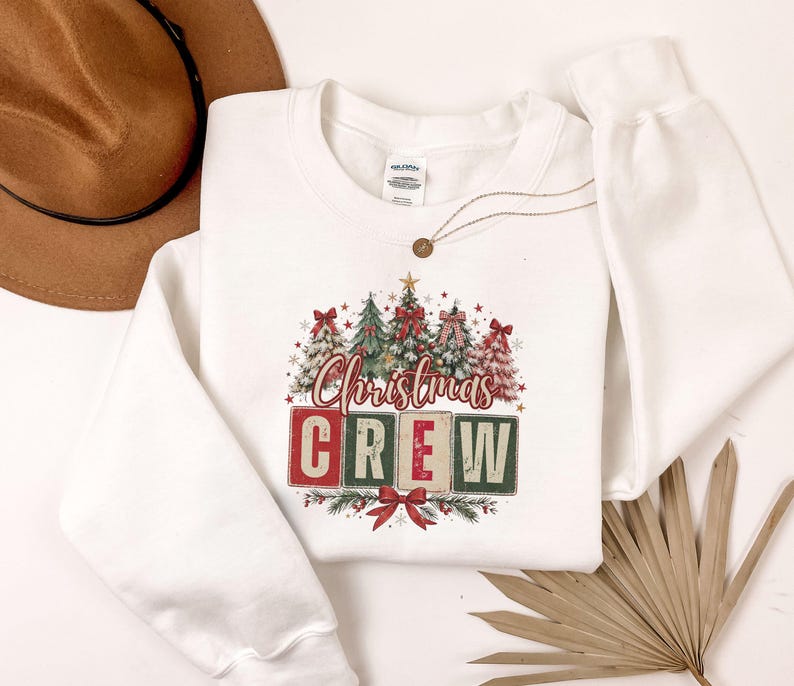 Op de afbeelding: Een wit sweatshirt met lange mouwen en een afbeelding van vier gestileerde kerstbomen versierd met rode strikken en sterren. Onder de bomen staat de tekst "Christmas CREW" in rode en groene blokletters met een rode strik onderaan.