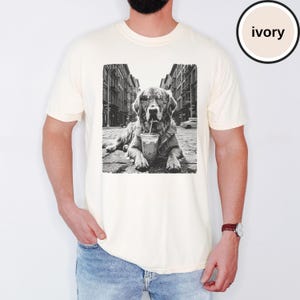 Peut inclure: Un t-shirt ivoire &agrave; manches courtes avec une impression graphique en noir et blanc repr&eacute;sentant un chien portant des lunettes de soleil et buvant dans un gobelet en plastique avec une paille, sur fond de rue urbaine. Col rond et coupe d&eacute;contract&eacute;e.