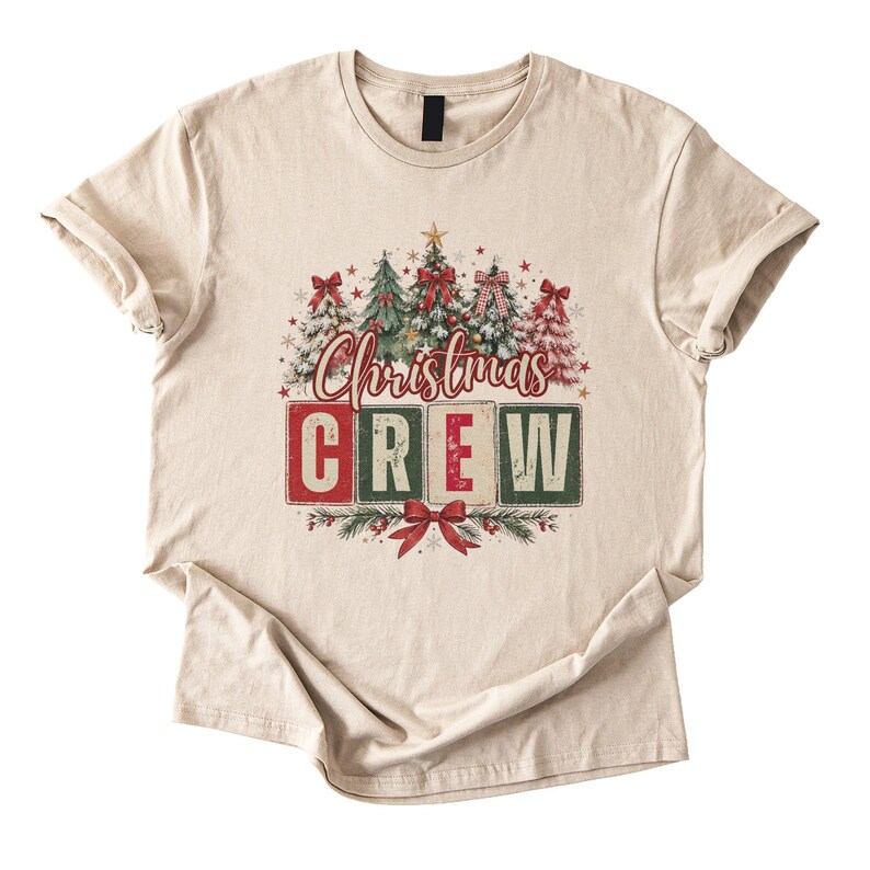Op de afbeelding: Een beige t-shirt met korte mouwen en een grafische print van kerstbomen met rode strikken en sterren. Onder de bomen staat de tekst "Christmas" in rode schrijfletters, gevolgd door het woord "CREW" in grote blokletters op vierkante tegels.