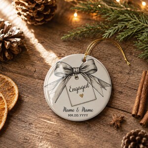 May include: A round white ceramic ornament featuring a black and white sketch of a bow tied to a gift tag. The tag displays the word Engaged with a small gold heart. Below the tag, the text Name & Name and MM.DD.YYYY is printed in black.