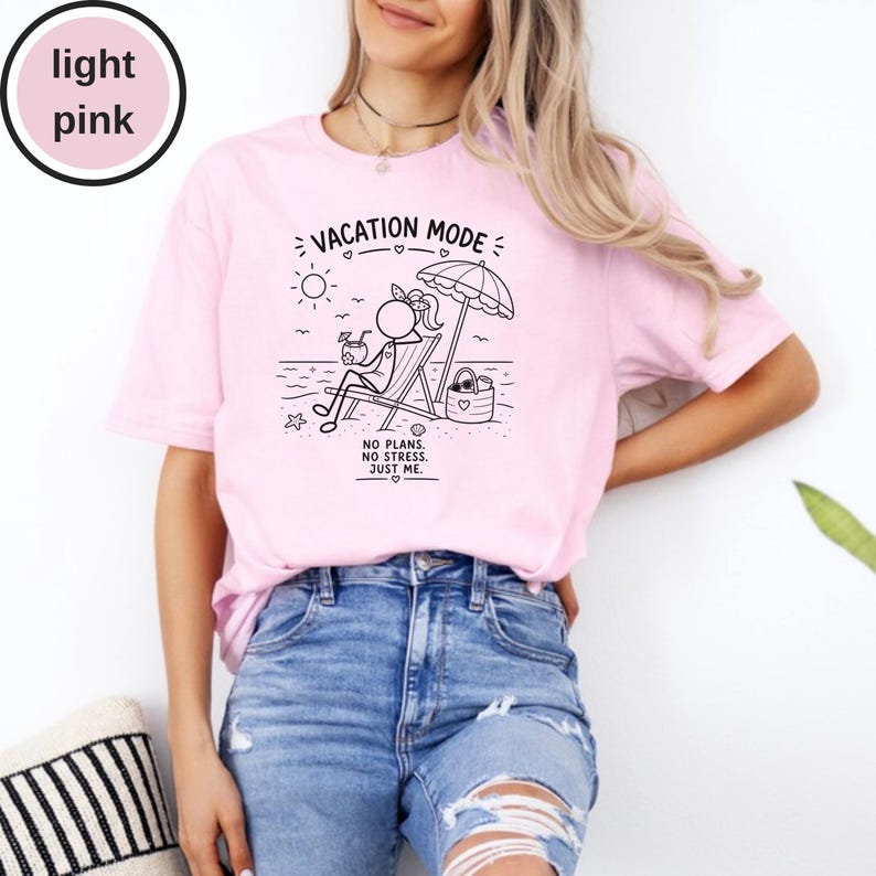 May include: Light pink t-shirt with a black graphic of a stick figure relaxing on a beach chair under an umbrella. The text "VACATION MODE" is above the image, and "NO PLANS. NO STRESS. JUST ME." is below. The shirt is made of soft material.