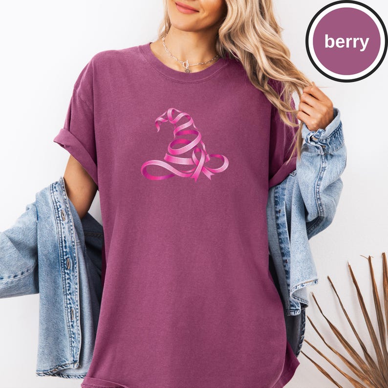 May include: A berry-colored short-sleeve t-shirt featuring a graphic of a witch hat constructed from a pink ribbon. The shirt has a crew neckline and a relaxed fit. A circular badge in the upper right corner contains the text berry.