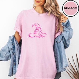 May include: A light pink t-shirt featuring a graphic of a witch hat constructed from a pink awareness ribbon. The shirt has short sleeves and a crew neckline. A circular badge in the upper right corner contains the text blossom in black.