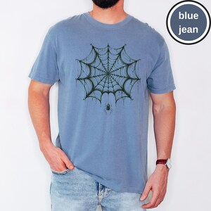 May include: A blue jean colored t-shirt featuring a dark green spider web graphic centered on the chest with a small spider hanging from the bottom of the web. The text "blue jean" is printed in white inside a circular badge in the top right corner.