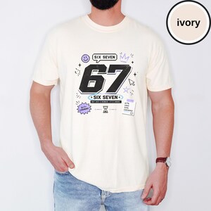 Pu&ograve; includere: Una t-shirt color avorio a maniche corte con una grande grafica nera del numero 67. Il design include il testo SIX SEVEN, una faccina sorridente, una corona, una stella, una clessidra e una nota con scritto 67% CHAOS 33% CONFIDENCE.