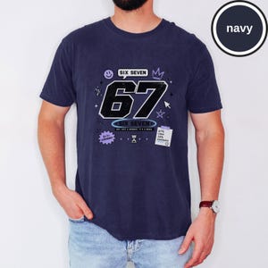 Pu&ograve; includere: Una t-shirt blu navy a maniche corte con una grande grafica del numero 67 in nero con contorni bianchi. Il design include piccole icone come una faccina sorridente, una corona, una stella, una clessidra e il testo SIX SEVEN, NOT JUST A NUMBER, IT'S A MOOD.
