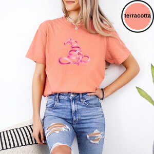 May include: A terracotta-colored short-sleeve t-shirt featuring a graphic of a witch hat formed from a pink ribbon. The shirt has a crew neckline and is paired with blue denim jeans that have distressed rips on the front of the legs.