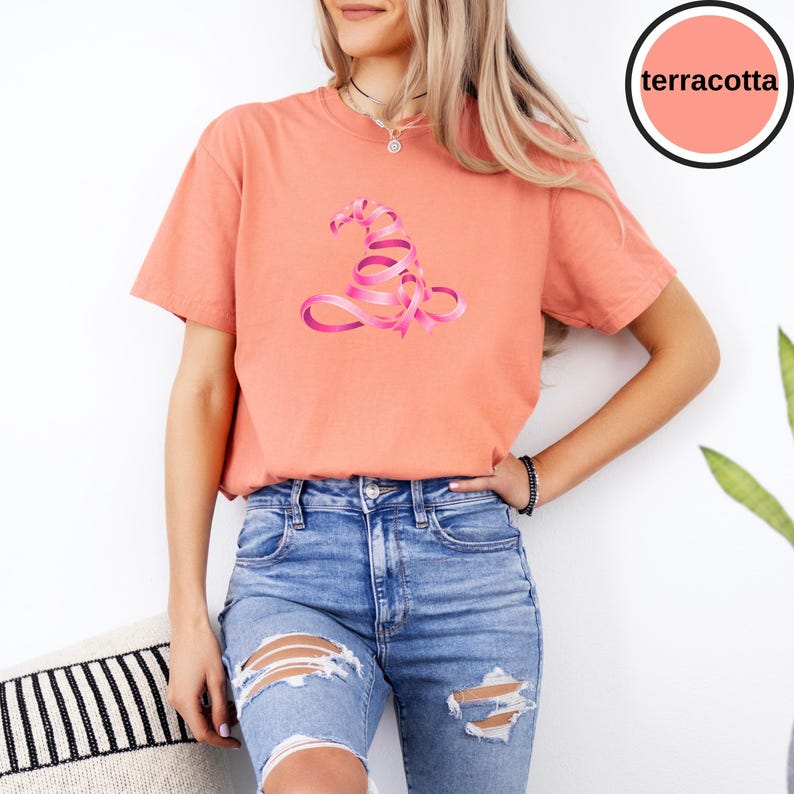 May include: A terracotta-colored short-sleeve t-shirt featuring a graphic of a witch hat formed from a pink ribbon. The shirt has a crew neckline and is paired with blue denim jeans that have distressed rips on the front of the legs.