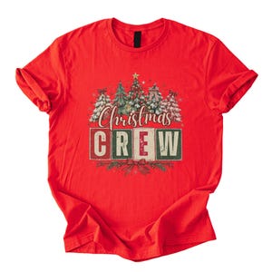 Op de afbeelding: Een felrood T-shirt met korte mouwen en een afbeelding van verschillende versierde kerstbomen boven de tekst "Christmas CREW" in blokletters. Het ontwerp bevat een rode strik aan de onderkant en kleine sterretjes tussen de bomen.