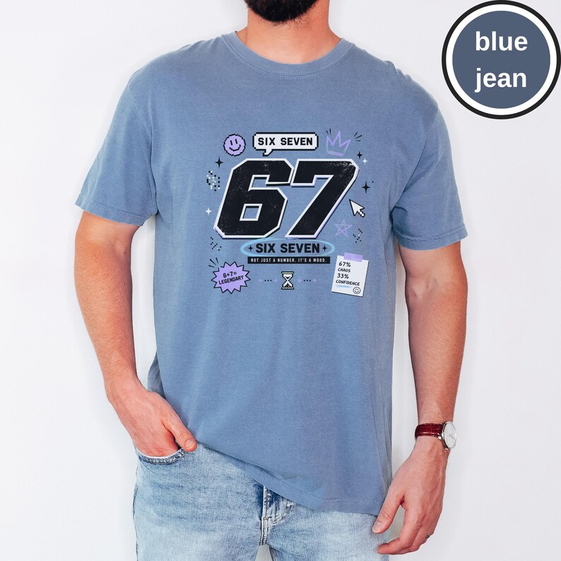 Pu&ograve; includere: Una t-shirt color blu jeans con una grande grafica del numero 67 in nero con contorni bianchi. Il design include il testo SIX SEVEN, 6+7=LEGENDARY e una piccola nota che riporta 67% CHAOS 33% CONFIDENCE, insieme a icone come smiley, corona e clessidra.