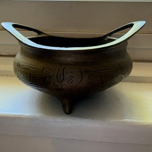 May include: A dark bronze-toned metal bowl featuring three small feet and two arched handles extending from the rim. The exterior surface of the bowl is decorated with etched linear patterns and circular motifs encircling the rounded body.