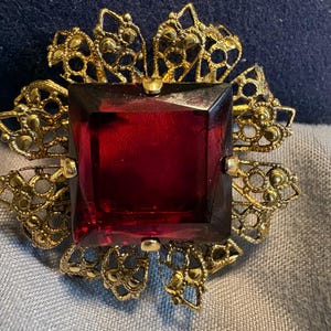 May include: A vintage brooch featuring a large, square, deep red gemstone at the center, surrounded by ornate gold-toned filigree. The filigree has a floral design, adding to the brooch's antique aesthetic. The brooch is set against a blue and gray background.