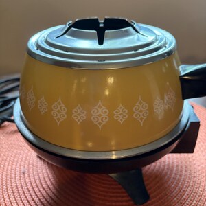 May include: A yellow electric fondue pot with a white repeating scroll pattern around the center. The pot features a silver metal rim at the top, a black base with three legs, and a black handle extending from the side with an attached power cord.