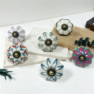 Peut inclure: Un ensemble de six boutons de tiroir en céramique en forme de fleur festonnée. Chaque bouton présente un motif peint à la main unique dans des tons de rose, bleu, rouge et vert, avec une pièce centrale en métal de couleur bronze.