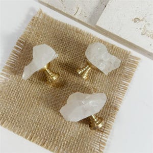 Peut inclure: Trois boutons de meuble avec des dessus en cristal de quartz naturel et des bases en laiton. Les boutons sont disposés sur une surface en toile de jute beige, avec un fond blanc et une dalle de pierre décorative.