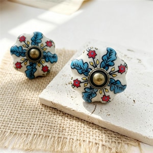 Puede incluir: Dos pomos de cajón de cerámica blanca con borde festoneado, con un patrón pintado a mano de hojas azules, pequeñas flores rojas y detalles amarillos. Cada pomo tiene una pieza central de metal en tono latón con un domo.
