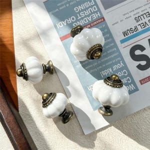 May include: Three white ceramic cabinet knobs with a fluted, pumpkin-like shape and antique brass-toned metal bases. Each knob features a decorative, scalloped metal collar where the ceramic meets the hardware, providing a classic, functional design.