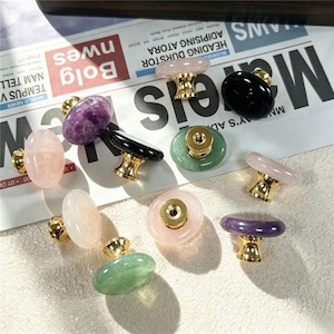 Puede incluir: Una colección de pomos redondos para armarios con tapas de piedras preciosas pulidas en tonos morado, rosa, verde, blanco y negro. Cada pomo está unido a una base de metal dorado brillante con un orificio roscado visible en la parte inferior.