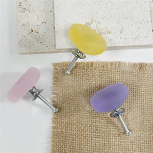 Peut inclure: Trois boutons de meuble colorés avec un aspect de verre de mer. Les boutons sont dans des tons de jaune, rose et violet, chacun avec une base et une vis argentées. Les boutons sont présentés sur une surface blanche avec une texture de toile de jute.