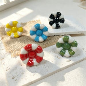 May include: Five ceramic drawer knobs shaped like flowers with alternating white and colored petals. The colors include yellow, blue, black, red, and green. Each knob features a glossy finish and a central circular button, providing a decorative accent.