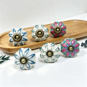 May include: Five ceramic drawer knobs with a flower-shaped design and a central brass-toned metal fixture. Two knobs feature blue painted petals, two feature pink and green petals, and one features green leaf patterns on a white background.