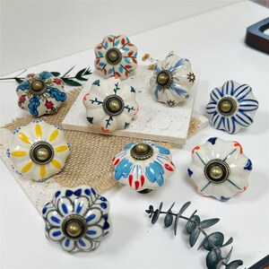 Puede incluir: Un conjunto de nueve pomos de cerámica para cajones con forma acanalada similar a una calabaza. Cada pomo presenta un patrón único pintado a mano en colores como azul, amarillo, rojo y verde, con un herraje central de metal color latón.