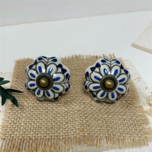 May include: Two round ceramic cabinet knobs featuring a white base with a blue and grey floral petal pattern. Each knob has a central circular brass-toned metal accent piece with a raised centre, providing a decorative finish to the hardware.