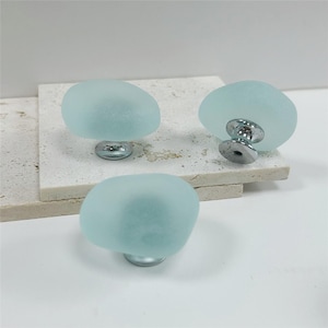 Peut inclure: Trois boutons de meuble vert d'eau avec une finition givrée et des bases chromées. Les boutons ont une forme arrondie et organique, ressemblant à des pierres lisses. Ils sont disposés sur une surface texturée de couleur claire.