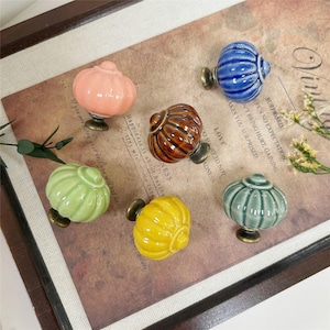 Peut inclure: Un ensemble de six boutons de tiroir en céramique de différentes couleurs, notamment rose, bleu, marron, vert clair, jaune et vert sauge. Chaque bouton présente une forme arrondie et nervurée rappelant une citrouille avec une base métallique.