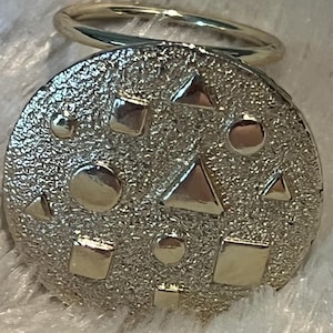 May include: A silver-toned compact mirror with a textured surface and geometric shapes. The mirror features squares, circles, and triangles. A silver ring is attached to the top of the compact.