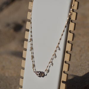 May include: A gold necklace with a seashell pendant, adorned with a speckled pattern. The chain is embellished with small pearls and gold beads. The necklace is displayed on a white stand with a wooden frame, set against a sandy background.