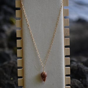 Op de afbeelding: Een goudkleurige ketting met een druppelvormige, roodbruine hanger. De ketting wordt gepresenteerd op een neutraal gekleurde sieradenstandaard met een houten frame. De achtergrond is wazig.
