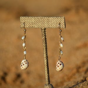 May include: Gold-toned dangle earrings featuring small white pearls and seashell charms. The earrings hang from delicate chains and are displayed on a neutral-toned jewelry stand. The seashells have brown spots.